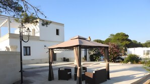 A covered outdoor seating area with chairs and a table, a white building in the background, and a small water feature in the foreground.
