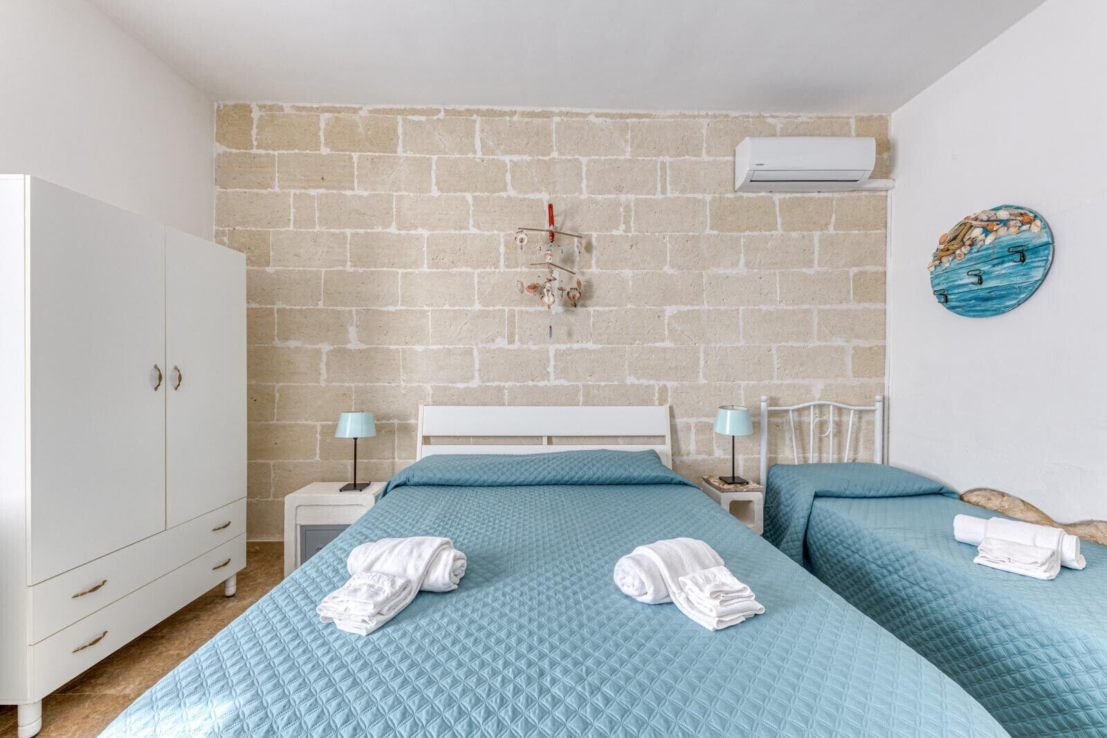 A bedroom with a brick wall, a white wardrobe, two beds with blue bedding, and a wall-mounted air conditioner.