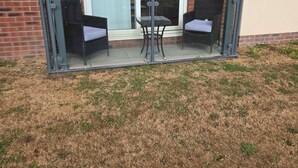 A balcony with two wicker chairs and a small table, a glass railing, and a brick wall with a door. - SeaBeach View Coastal Path Ground Floor Apartment (LLANELLI)