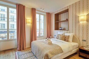 Ein Hotelzimmer mit einem großen Bett, einem Nachttisch, Vorhängen an einem Fenster und einem Wandregal mit einem Buch.