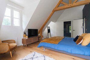 A bedroom with a sloped ceiling, a bed with a blue and brown blanket, a wooden dresser, a television, and a chair. - Exceptional House with Pool 12P – Englesqueville (Englesqueville-en-Auge)