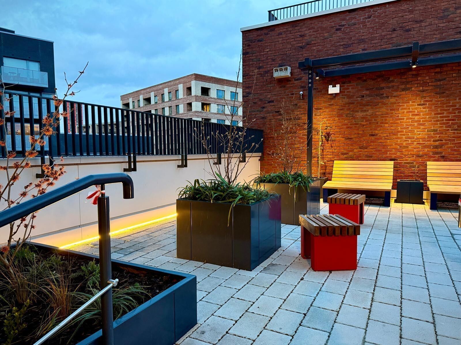 Een terras met plantenbakken en een bank, een bakstenen muur en een modern gebouw op de achtergrond.