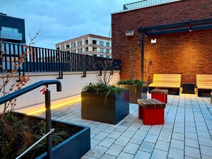 An outdoor seating area with planters and a bench, a brick wall, and a modern building in the background. - Student Only Ark Canning Town (London)
