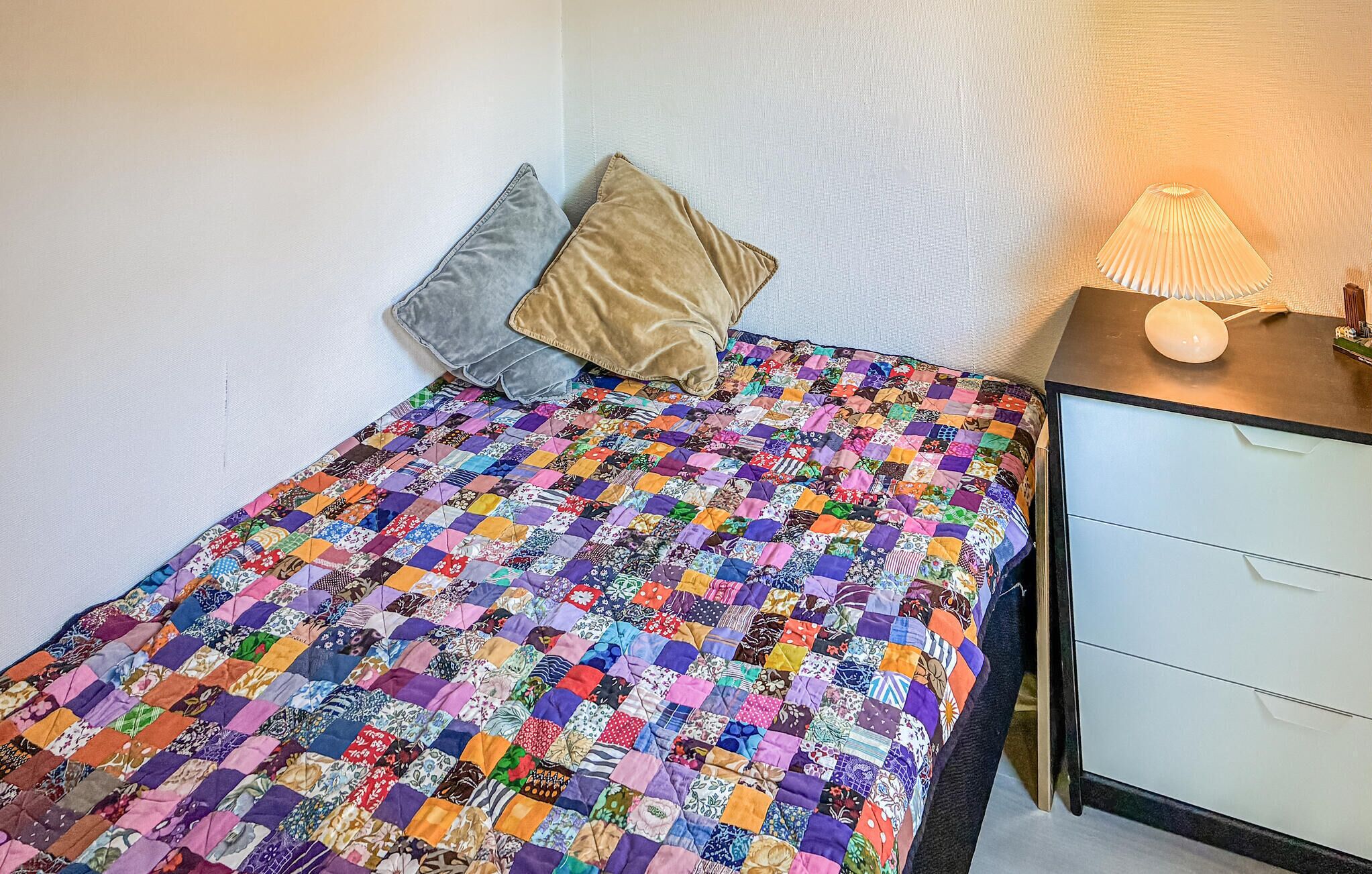 A bed with a colorful patchwork quilt, two pillows, a bedside table with a lamp, and a white nightstand with drawers.