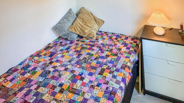 A bed with a colorful patchwork quilt, two pillows, a bedside table with a lamp, and a white nightstand with drawers.