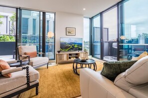 Ein modernes Wohnzimmer mit einer Couch, Sesseln, einem Couchtisch, einem Fernseher und einem großen Fenster mit Blick ins Freie.
