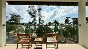A balcony with two wooden chairs and a small table, overlooking a beach with trees and a clear sky. - Boutique 1BR Suite with Sea View, Patong (Tambon Patong)