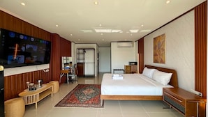 A hotel room with a large flat-screen TV, a bed with white linens, a wooden nightstand, a desk with a chair, and a patterned rug. - Deluxe Double Room with Sea View Balcony (Tambon Patong)