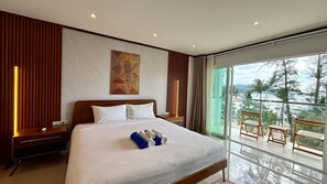A hotel room with a large bed, a nightstand, a bedside table, a window with a view of the outdoors, and a balcony with wooden furniture. - Deluxe Double Room with Sea View Balcony (Tambon Patong)