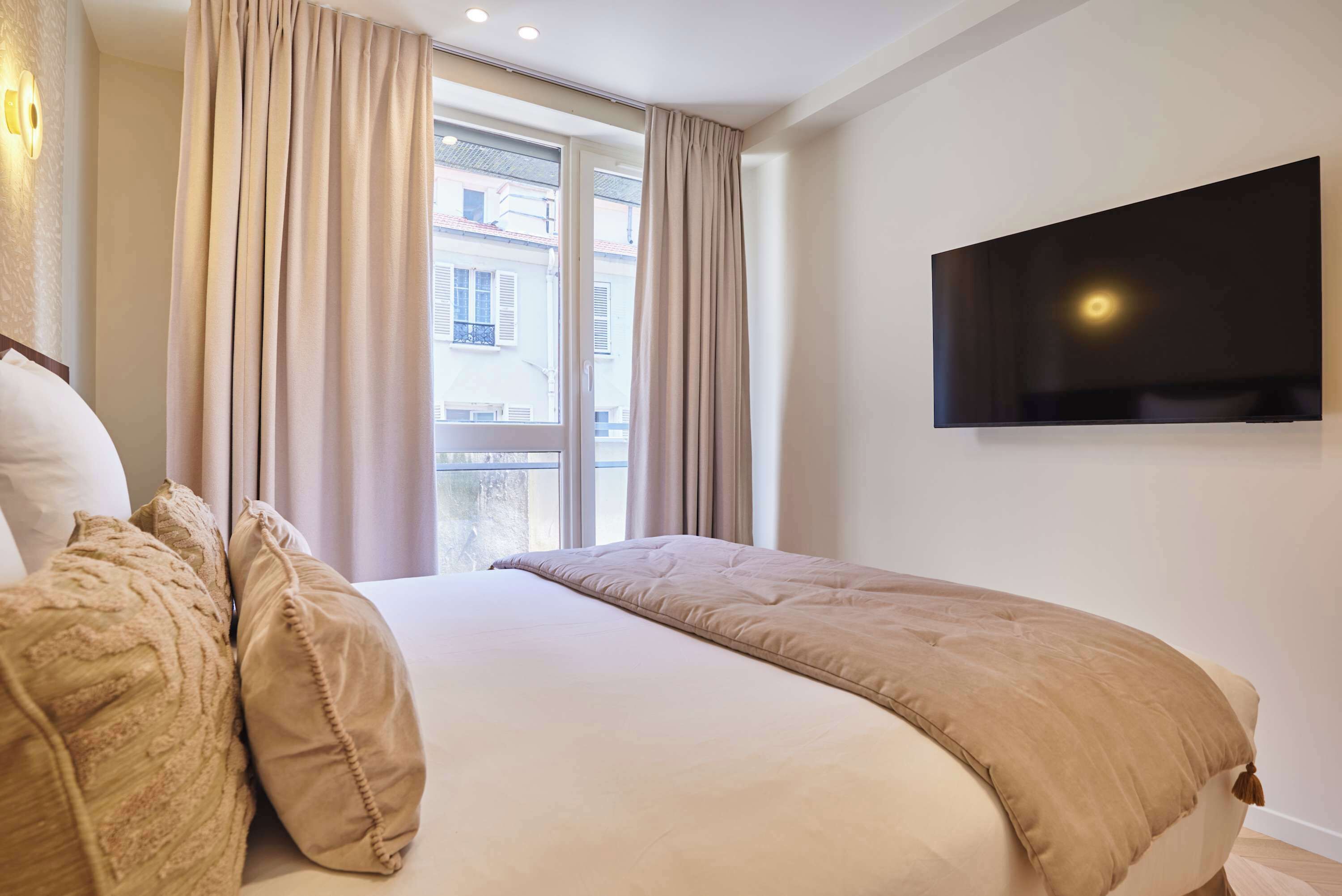 Una camera d'albergo con un letto, tende, una TV fissata al muro e una finestra vista edifici.
