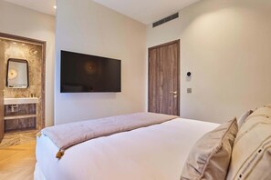 Ein Schlafzimmer mit Bett, einem an der Wand befestigten Fernseher, einer Holztür sowie einem Badezimmer mit Spiegel und Waschbecken.