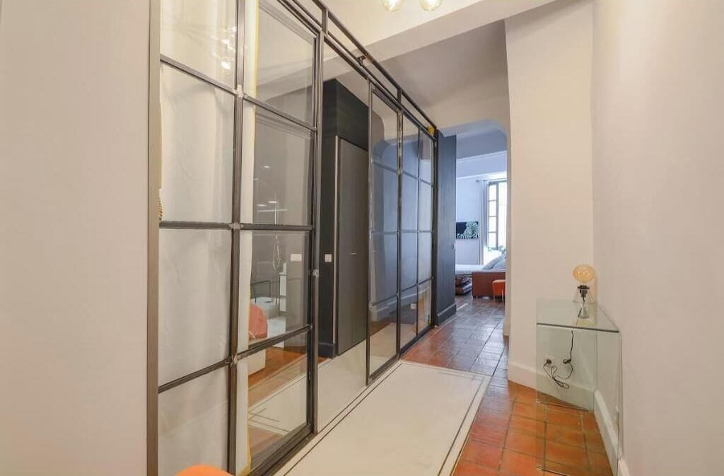 A hallway with a glass partition, a glass cabinet with a light, and a tiled floor.