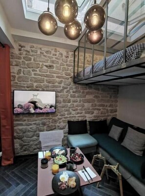 A cozy room with a stone wall, a green sofa, a dining table with breakfast, and a hanging light fixture. - The Little Workshop of Light • Air Conditioning & Sunrooms in the Heart of Paris (Île-de-France)