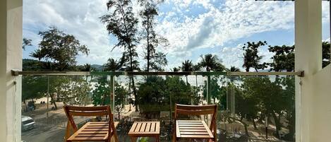 A balcony with two wooden chairs and a small table, overlooking a beach with trees and a clear sky.