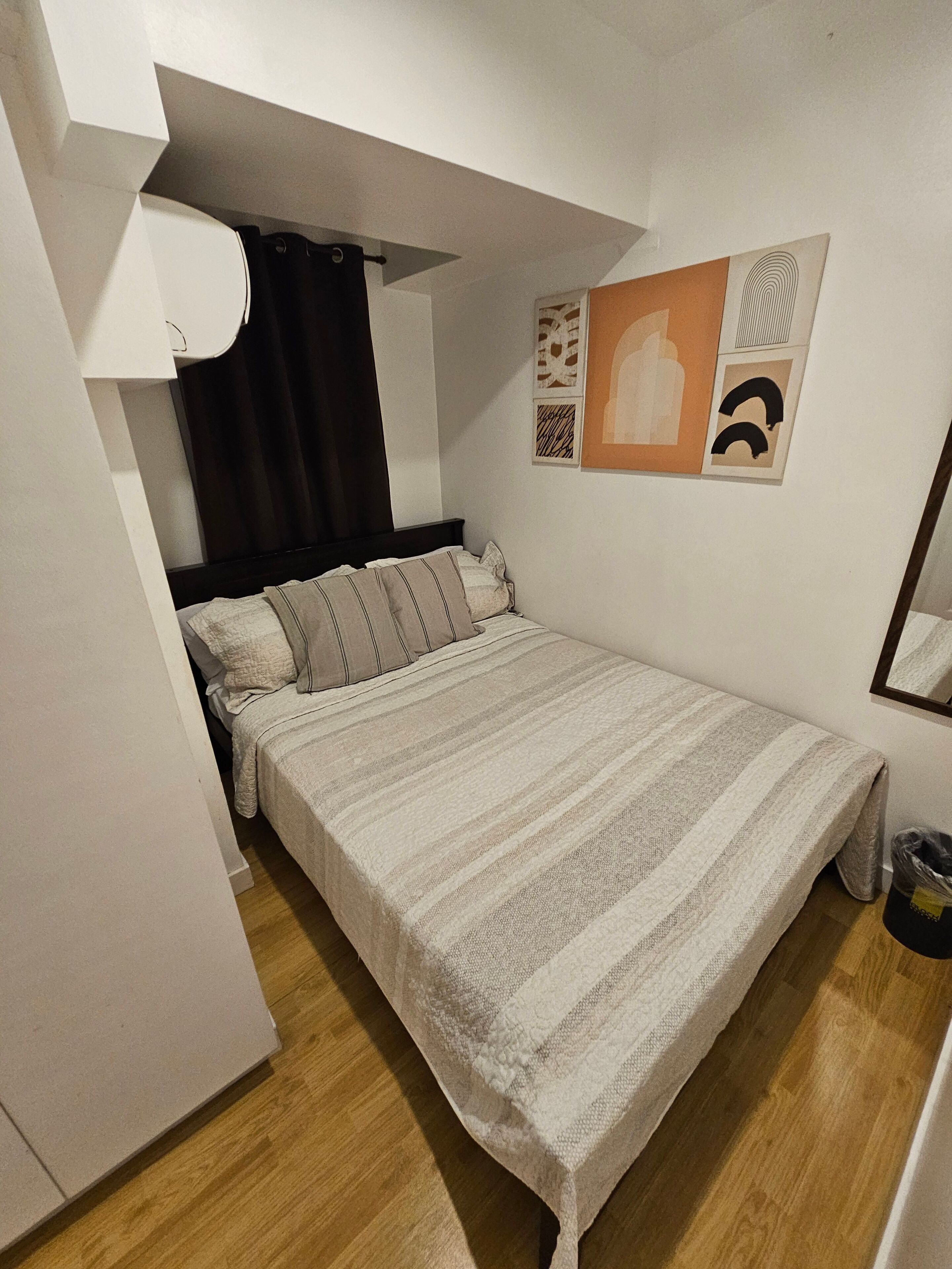 A bedroom with a bed, a window with curtains, a wall-mounted artwork, and a trash bin.