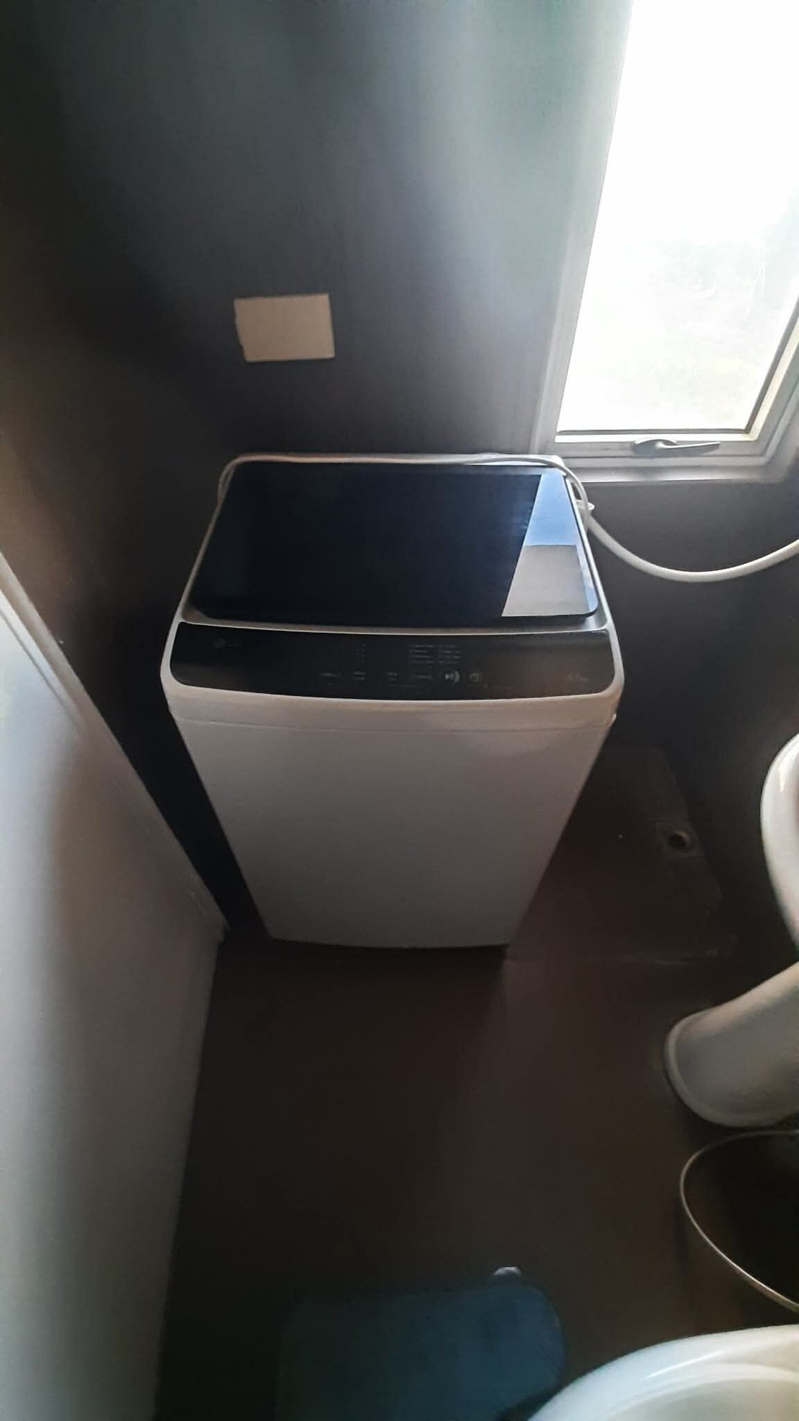 Une petite machine à laver blanche, placée dans une petite pièce qui comprend une fenêtre et des toilettes.