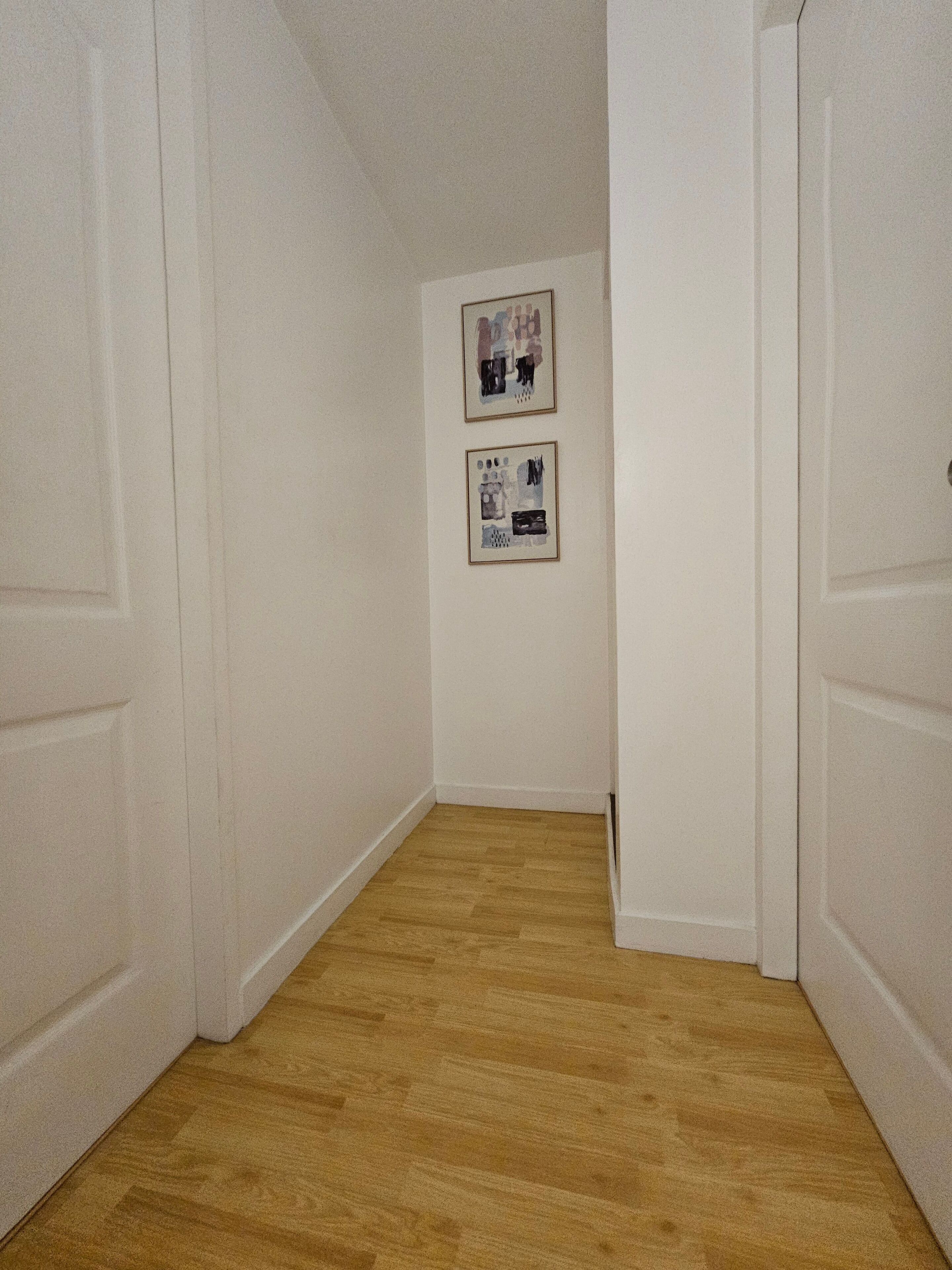 A narrow hallway with wooden flooring and white walls. There are two framed pictures on the wall.
