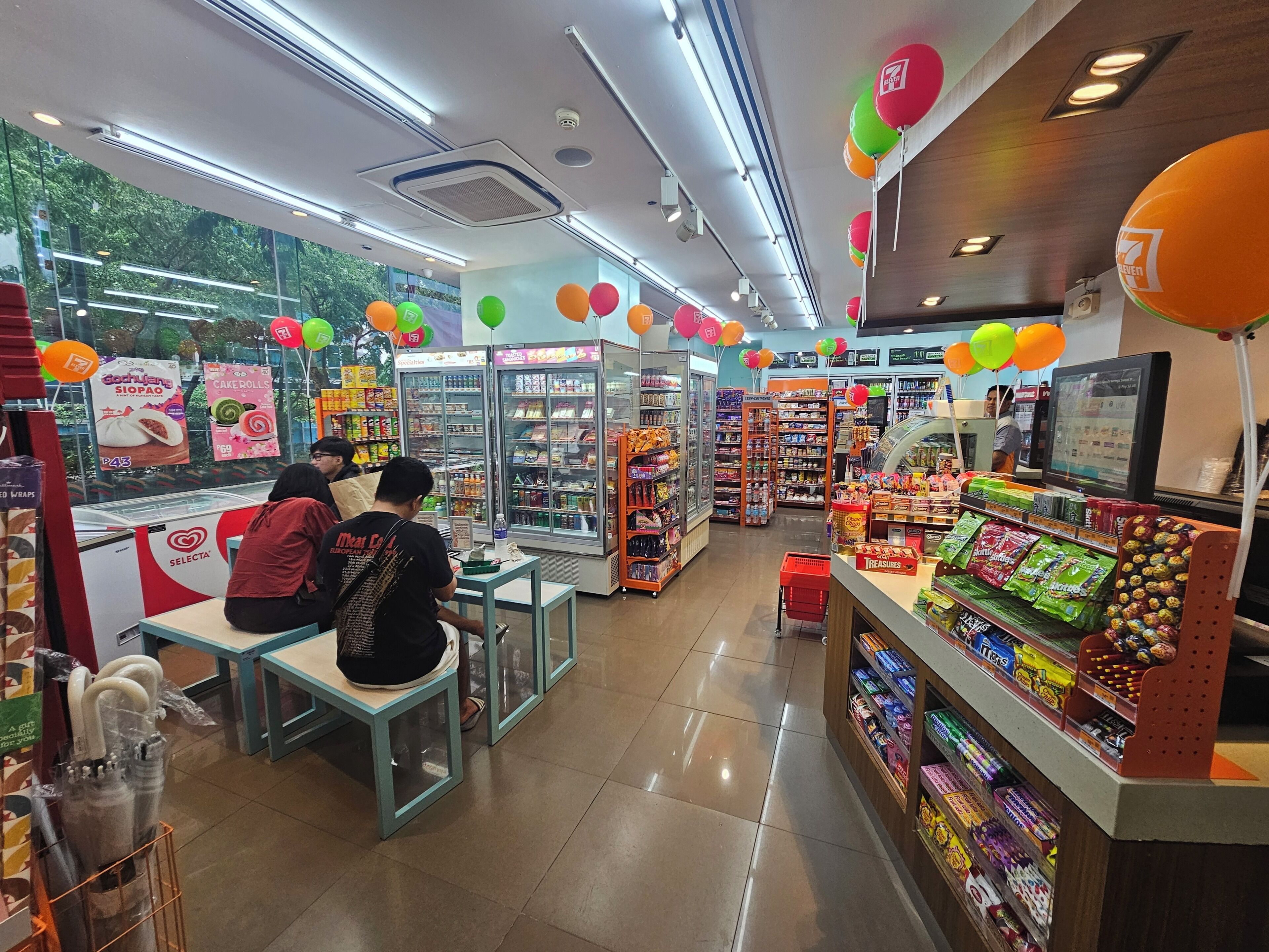 A brightly lit convenience store with refrigerated sections, shelves stocked with snacks, and a checkout area.