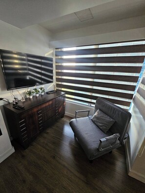 Ein Zimmer mit dunklem Holzboden, einem grauen Sofa, einem an der Wand befestigten Fernseher und einem Fenster mit Rollläden.