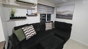 Un soggiorno con un divano nero, un condizionatore d'aria a parete, una finestra con vista e un quadro incorniciato appeso al muro.