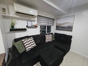 Ein Wohnzimmer mit schwarzem Sofa, einer Wand-Klimaanlage, einem Fenster mit Ausblick und einem gerahmten Bild an der Wand.