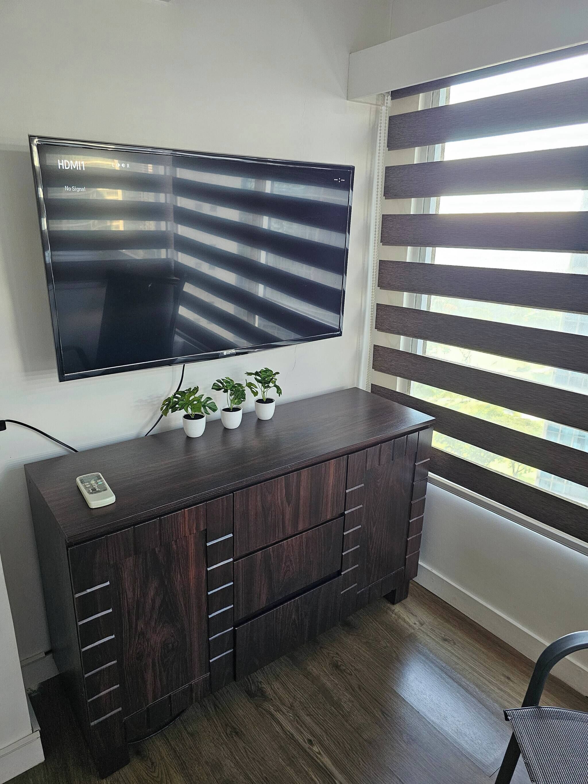 A flat-screen TV mounted on a wall, a dark wooden cabinet with drawers, and a window with horizontal blinds.