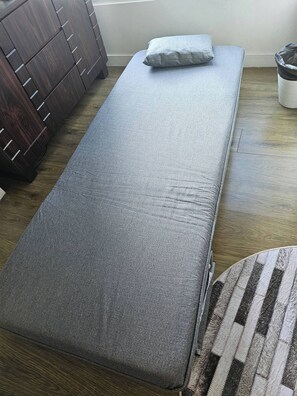 Ein Zimmer mit einem Einzelbett, einem Holzschrank und einem Mülleimer.