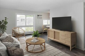 Ein Wohnzimmer mit einer Couch, einem runden Couchtisch, einem Fernseher auf einer Holzkommode und einem großen Fenster mit Jalousien.