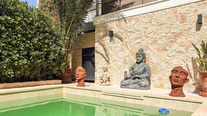 Área de piscina com estátua de Buda e estátua infantil, cercada por vegetação e um muro de pedra.