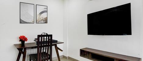 Una habitación con una mesa de comedor, una silla, un televisor montado en la pared y un estante pequeño debajo del televisor.