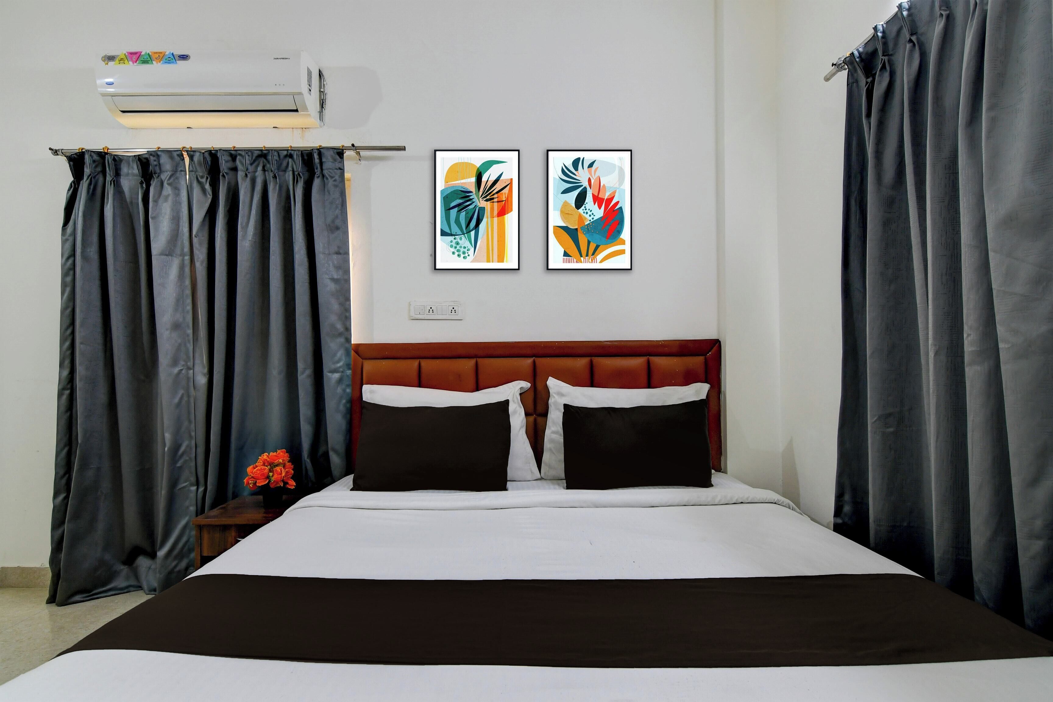 Habitación de hotel con una cama, dos cuadros enmarcados, una mesita de noche con un florero y un aire acondicionado instalado en la pared.