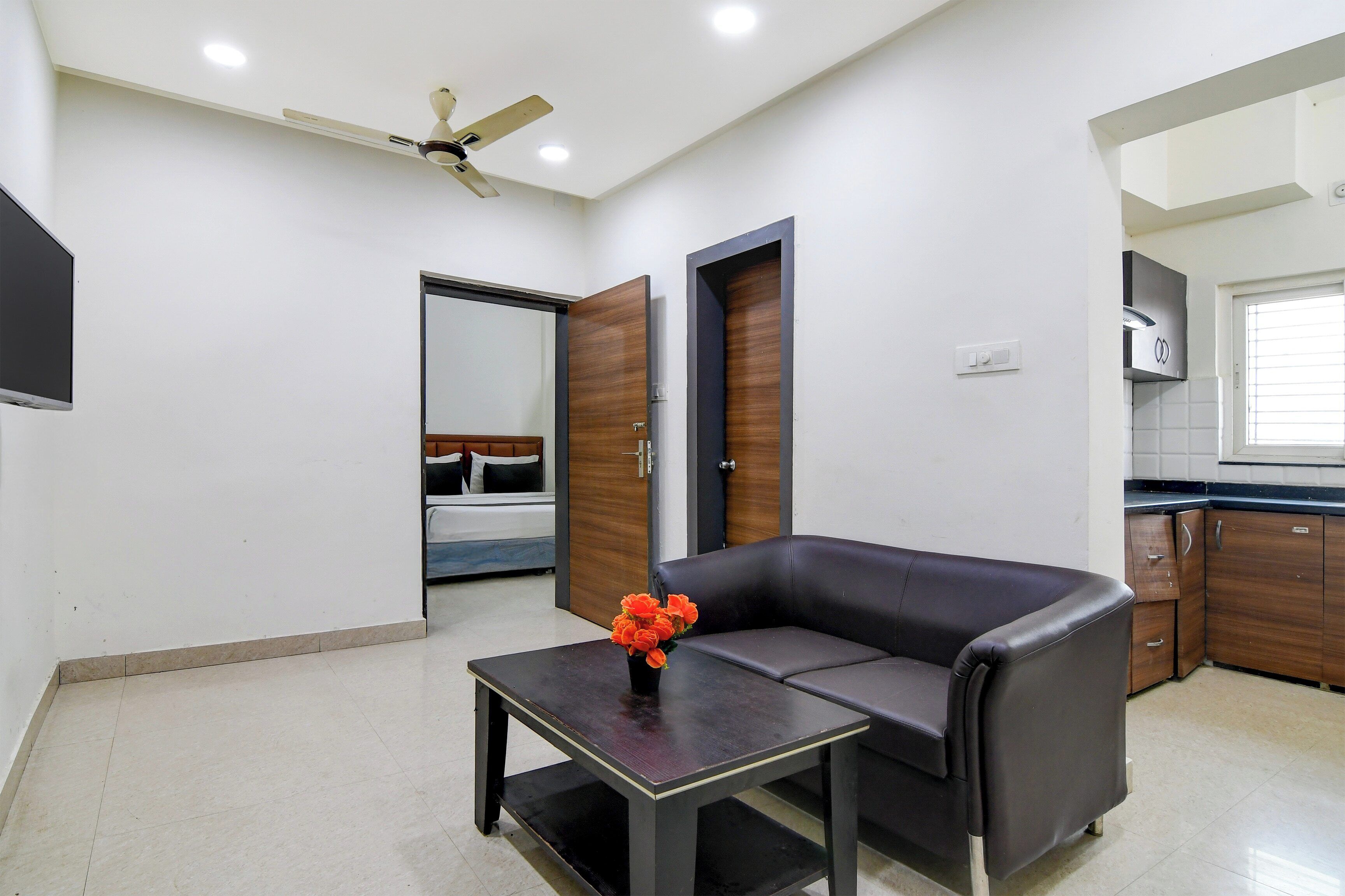 Una sala de estar moderna con un sofá, una mesita con un florero, un ventilador de techo y una zona de cocina con armarios y una ventana.