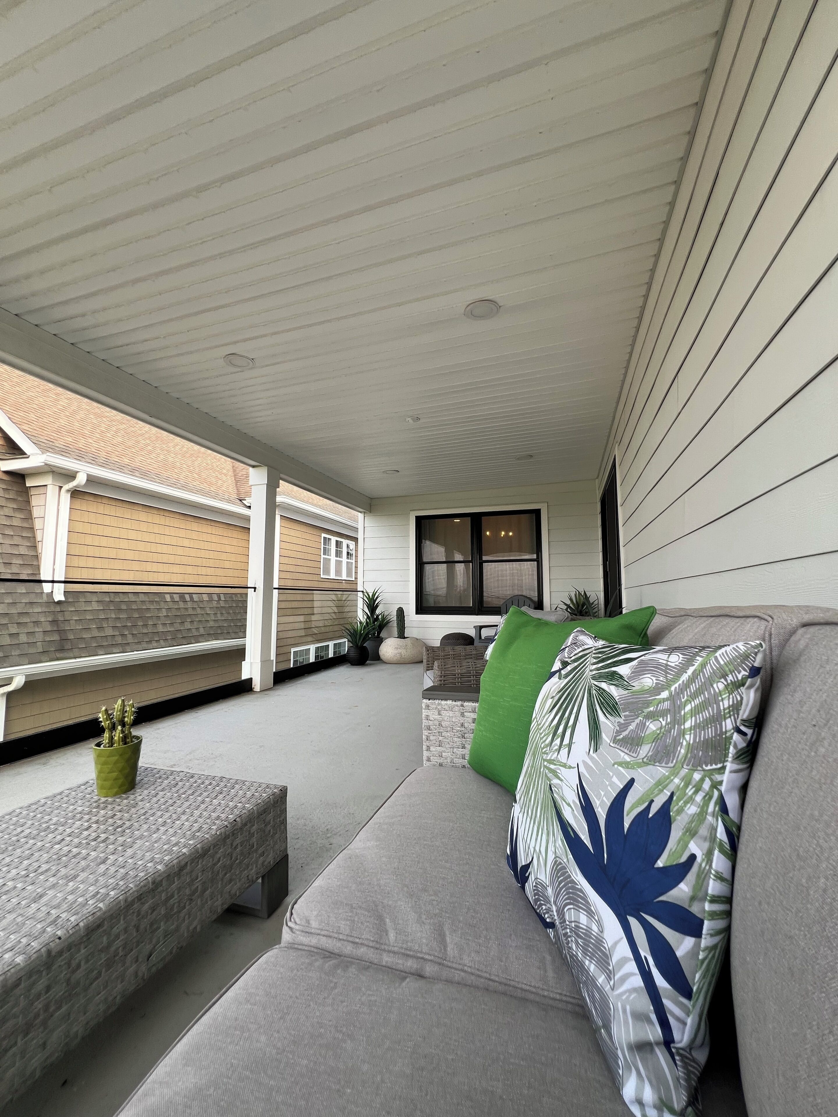 A covered patio area with a cushioned seating arrangement, a potted plant, and a window with blinds.