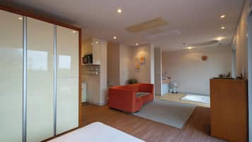 A modern hotel room with a bed, a red sofa, a small kitchen area, and a bathroom.