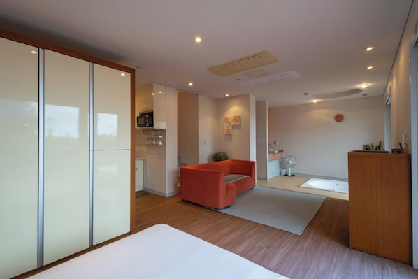 A modern hotel room with a bed, a red sofa, a small kitchen area, and a bathroom.