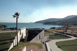 A modern building with a sloped roof and a concrete facade, situated near a coastal area with a clear view of the sea and distant hills. - Tropical Dream Resort (Geoje)