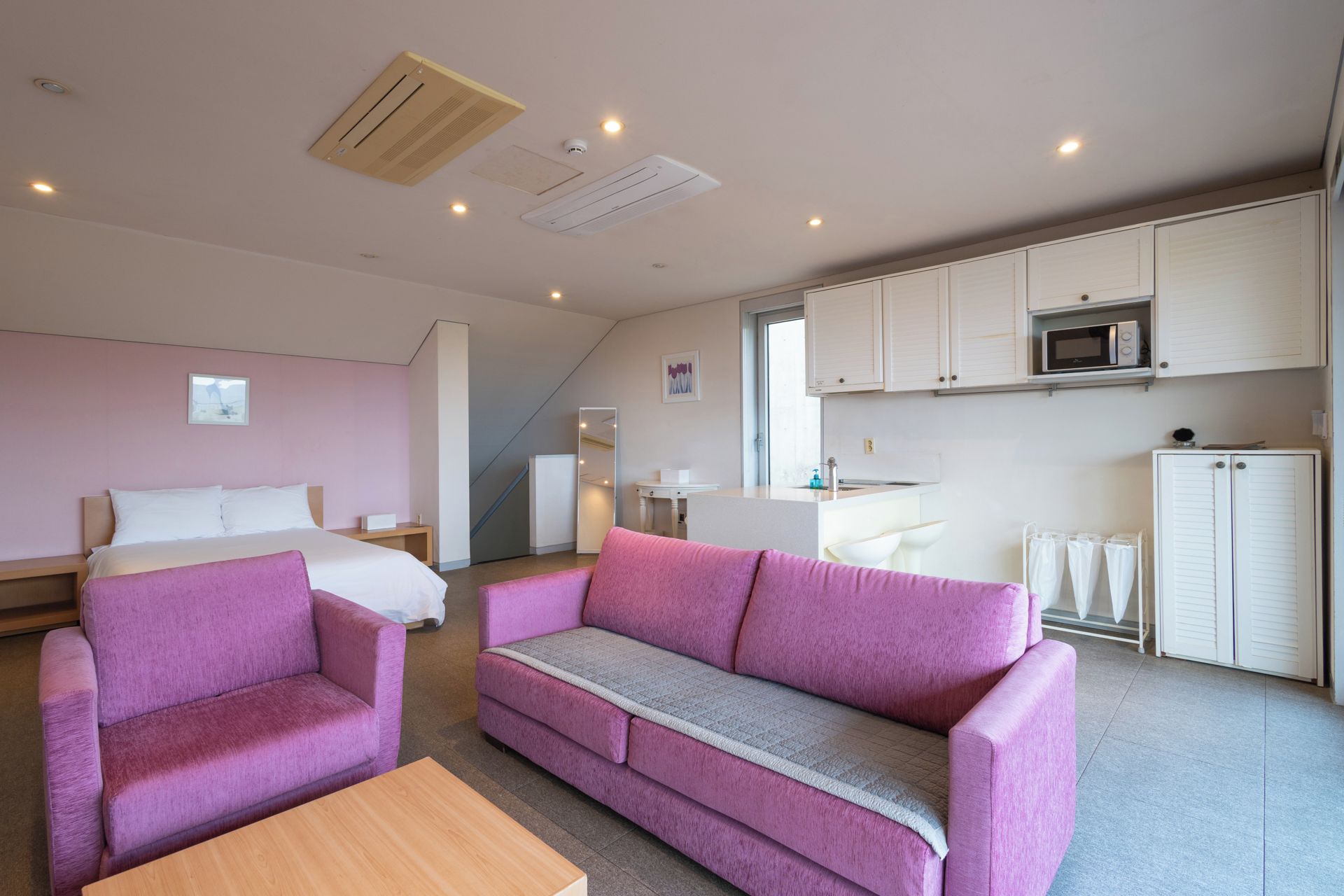 Habitación de hotel moderna con cama, sofá rosa, una mesa de centro de madera, kitchenette y baño con ducha.