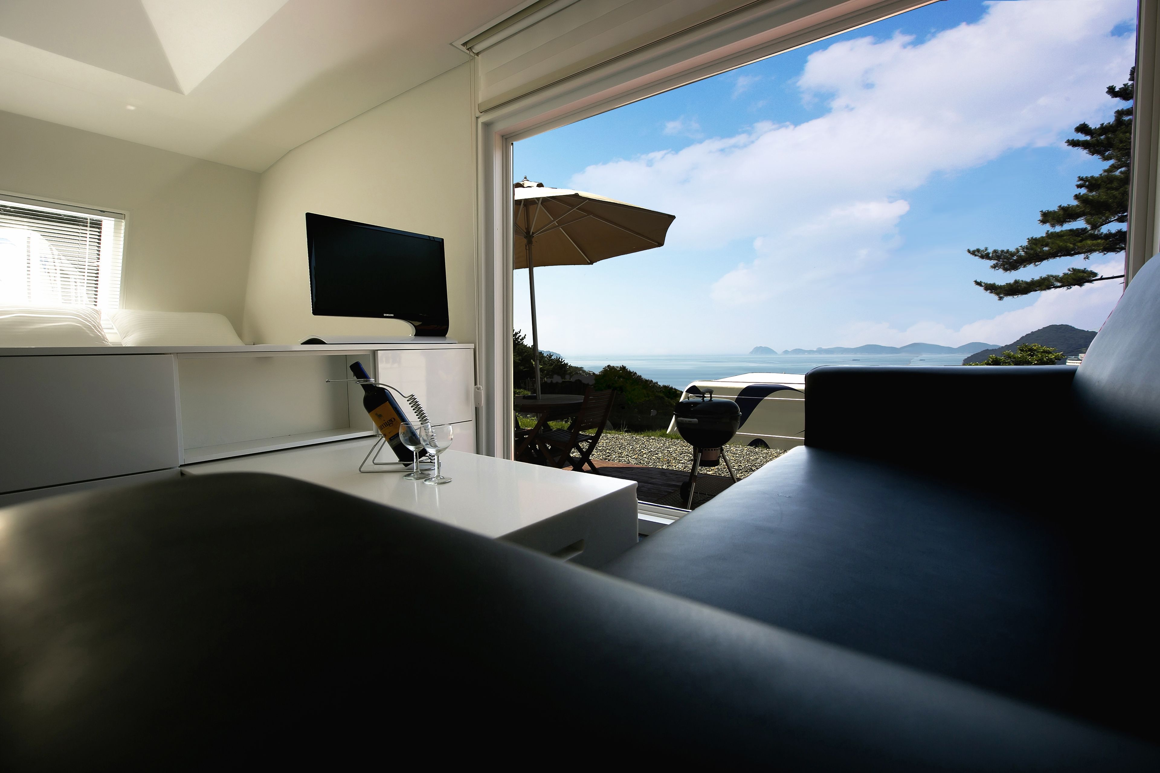 A modern bedroom with a TV on the wall, a bedside table with a bottle and glasses, and a view of the sea through a large window.