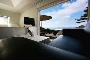 A modern bedroom with a TV on the wall, a bedside table with a bottle and glasses, and a view of the sea through a large window.
