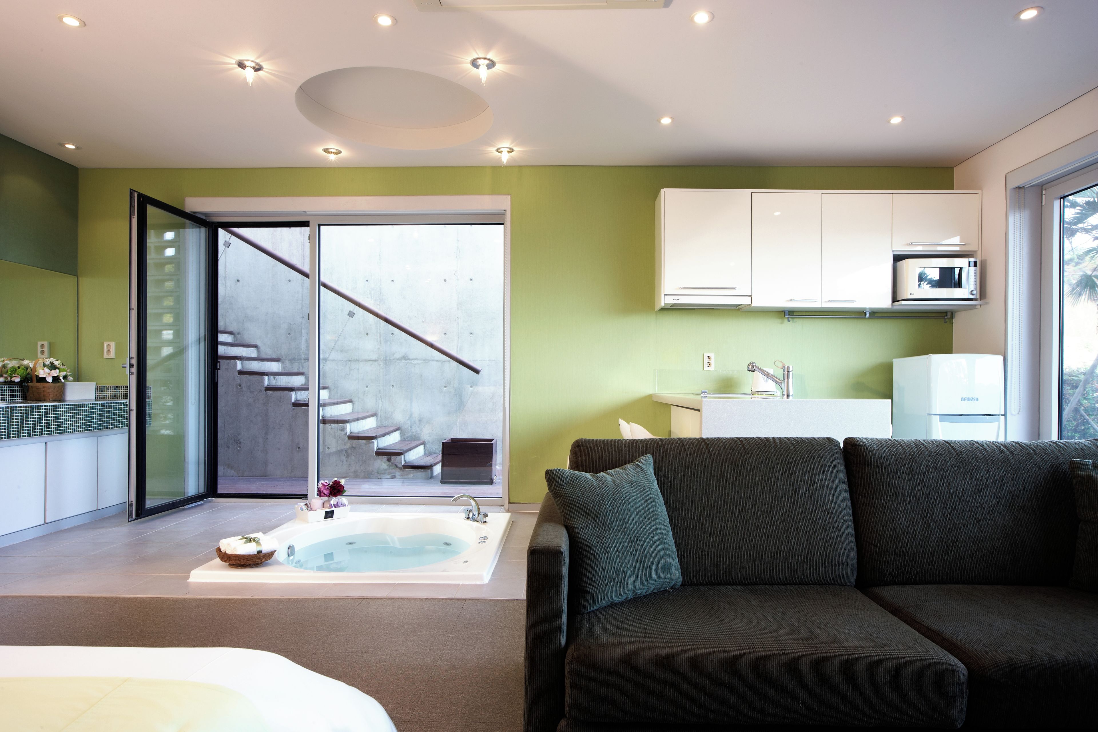A modern interior with a glass door leading to a staircase, a seating area with a dark sofa, a built-in kitchen with a microwave, and a bathtub.