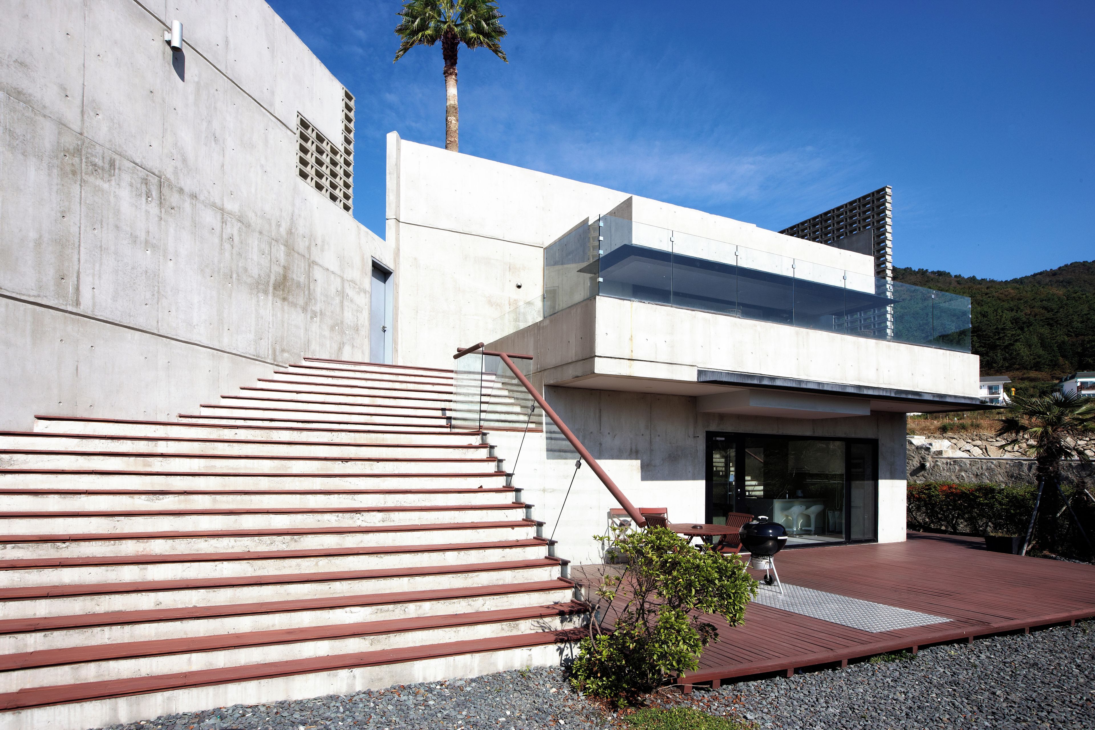 Un edificio moderno con fachada de hormigón, una extensión de vidrio y una escalera que lleva a un espacio exterior cubierto con mesa y sillas.