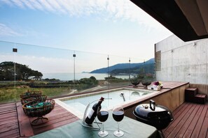 A rooftop terrace with a glass-enclosed hot tub, a barbecue grill, and a table with a bottle of wine and glasses.