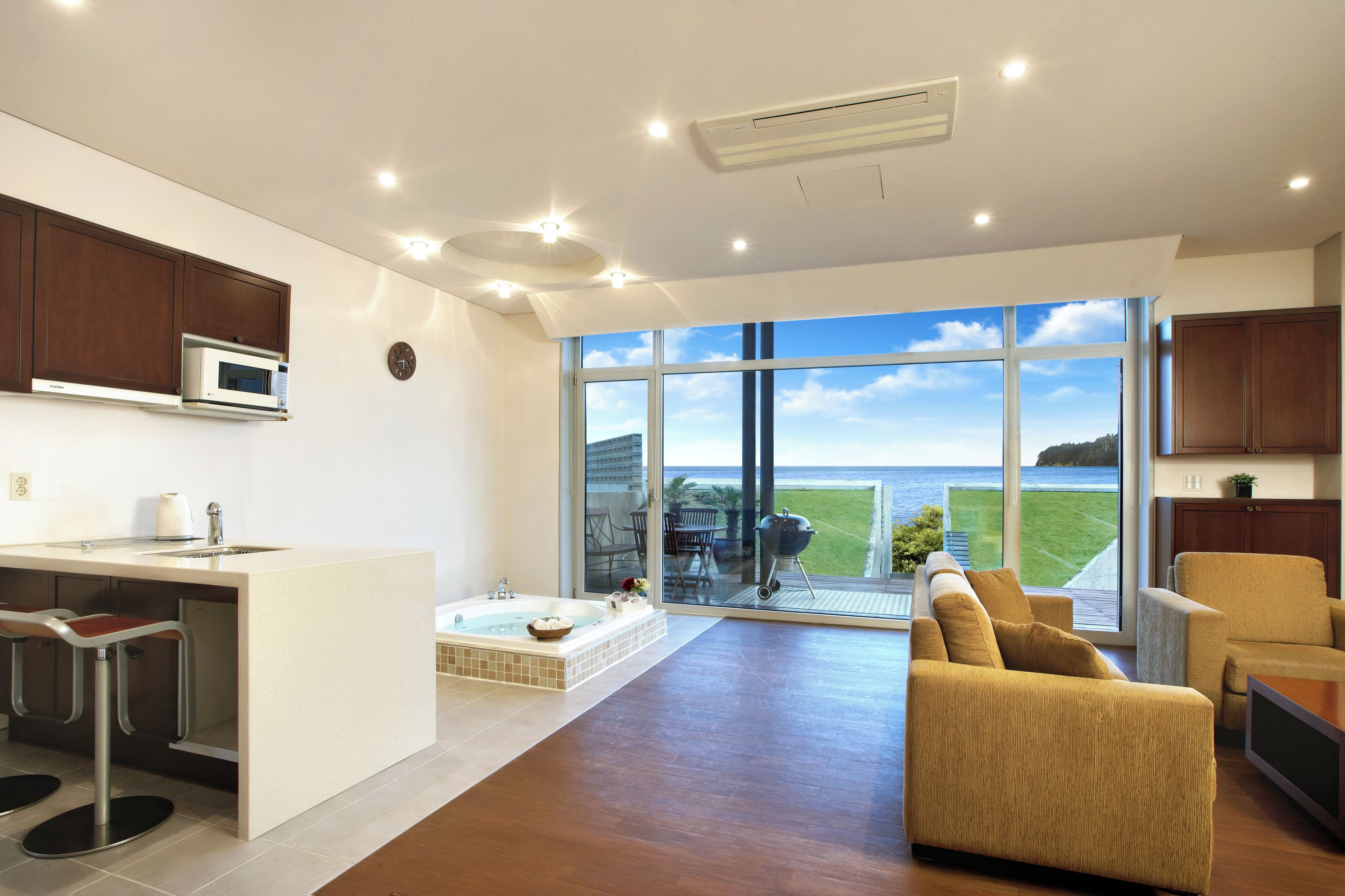 A modern kitchen with a built-in microwave, a dining area with a bar stool, a large window with a view of the sea, and a cozy seating area with a sofa and armchair.