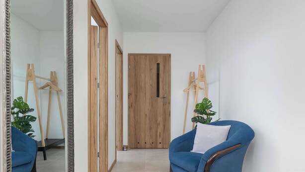 Ein Flur mit einer Holztür, einem blauen Sessel mit weißem Kissen und einem Spiegel, der den Raum widerspiegelt.