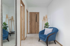 Ein Flur mit einer Holztür, einem blauen Sessel mit weißem Kissen und einem Spiegel, der den Raum widerspiegelt.