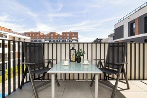 Eine Terrasse im Freien mit einem Glastisch und zwei Stühlen, einem Blumentopf und Blick auf moderne Gebäude im Hintergrund.