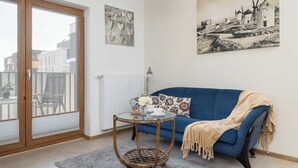 Ein Zimmer mit einem blauen Sofa, einem Glastisch mit einer Vase, einem Korbstuhl und einem großen Fenster mit Blick auf Gebäude.