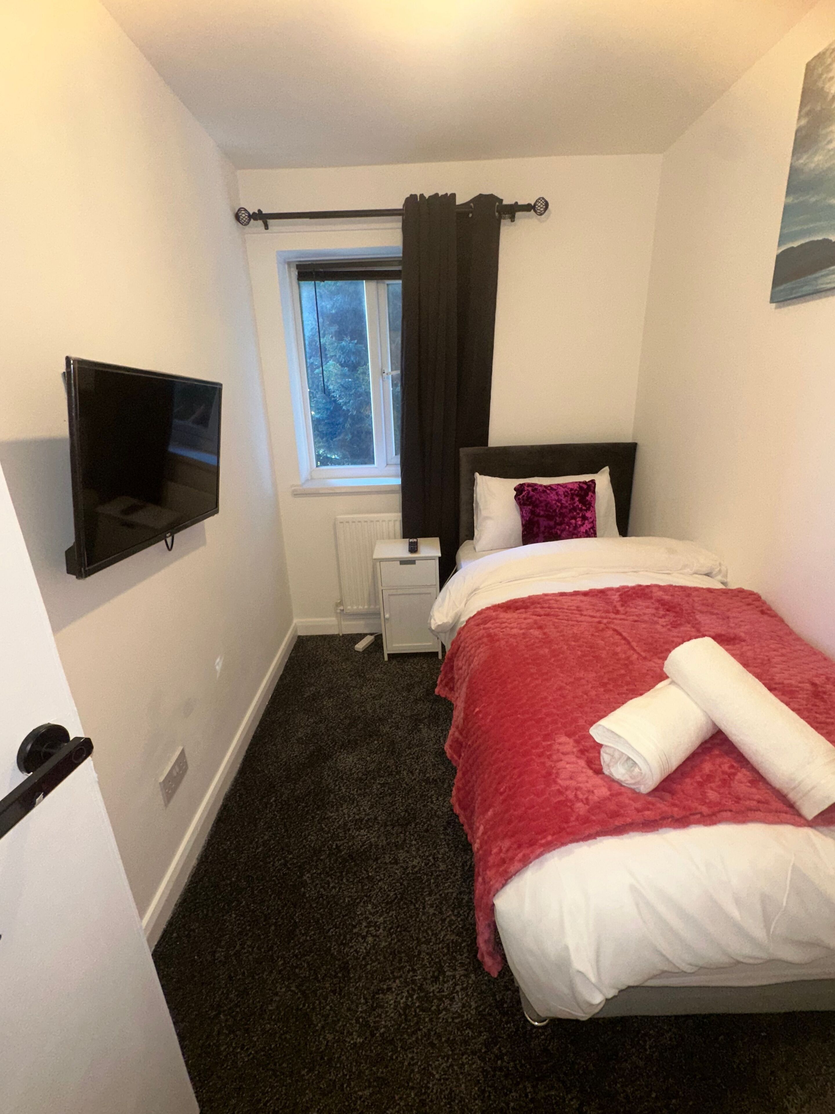 A small, neatly arranged bedroom with a bed, a television mounted on the wall, a window with curtains, and a radiator.