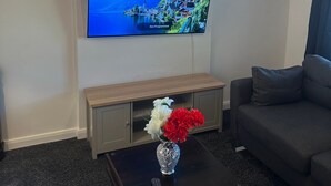 Een woonkamer met een flatscreen televisie aan de muur, een houten salontafel met een bloevas en een grijze bank.