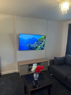Una sala de estar con un televisor de pantalla plana montado en la pared, una mesa de centro de madera con un jarrón de flores y un sofá gris.